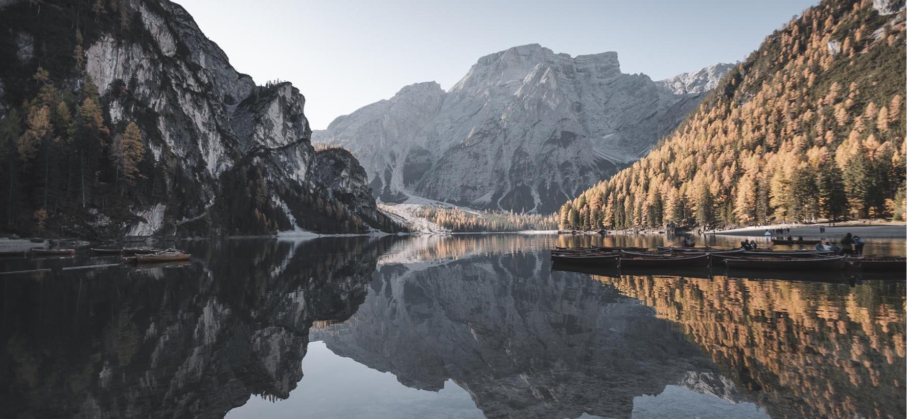 Ruhiger Bergsee in den Dolomiten mit Spiegelung der umliegenden Gebirge, bewaldeten Hänge und herbstlich gefärbter Landschaft.