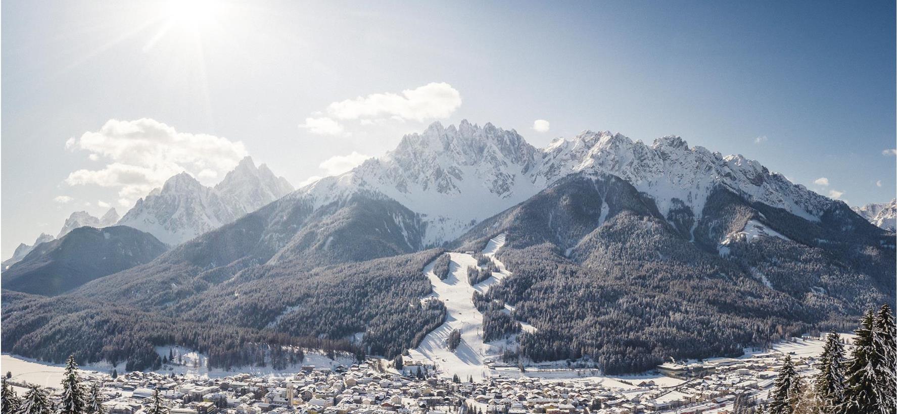 San Candido in inverno con tetti innevati, alberi e montagne sullo sfondo.