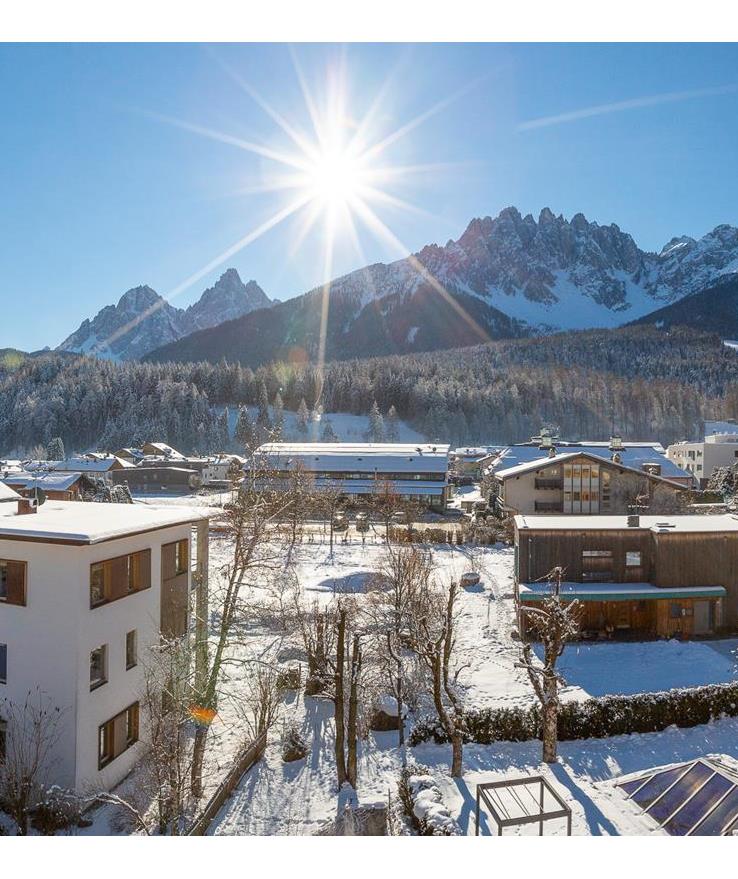 Vista panoramica sulle Dolomiti con paesaggio montano innevato, abitazioni e splendido sole.
