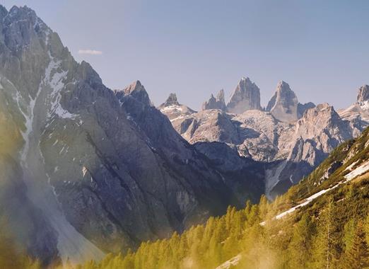 Paesaggio montano alpino nelle Dolomiti con prati verdi in primo piano, imponenti cime rocciose e montagne sullo sfondo.