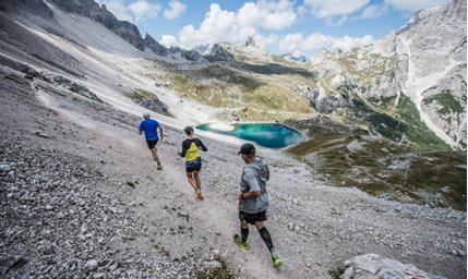 Three mountain runners on steep terrain, surrounded by an imposing mountain landscape with an alpine lake in the background.