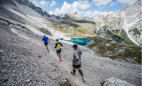 Three mountain runners on steep terrain, surrounded by an imposing mountain landscape with an alpine lake in the background.