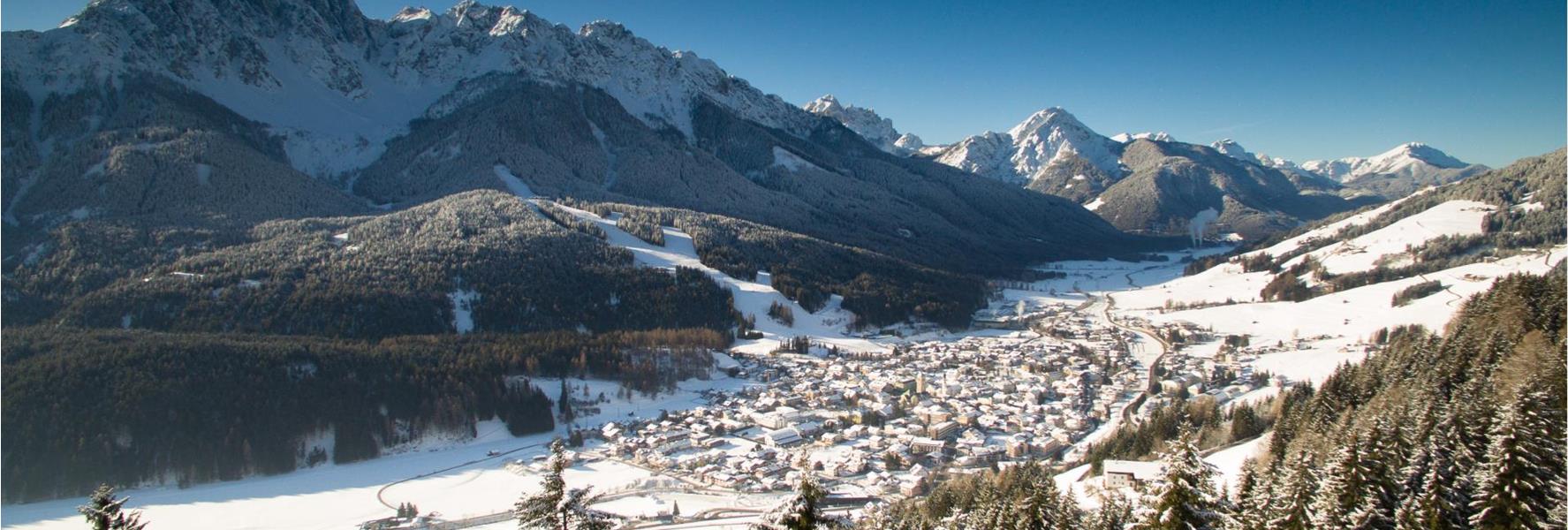 Winter view of San Candido with snow-covered forests and the Baranci mountain group in the Dolomites in the background.