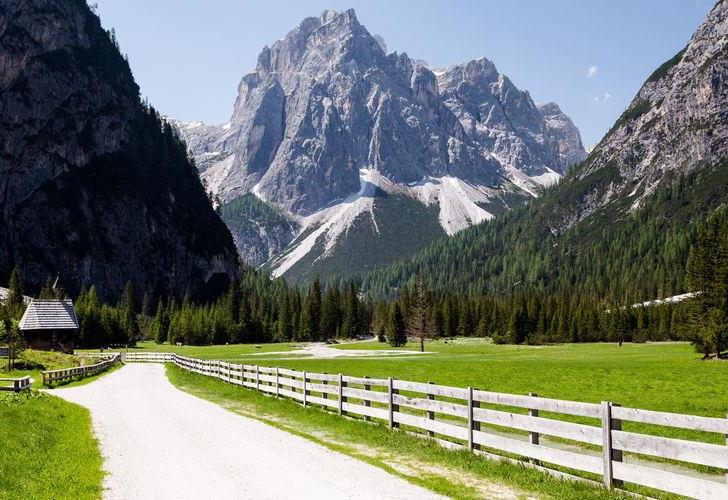 Ein breiter Wanderweg durch ein grünes Tal in den Dolomiten mit Wiesen, Wäldern und markanten Berggipfeln im Hintergrund.