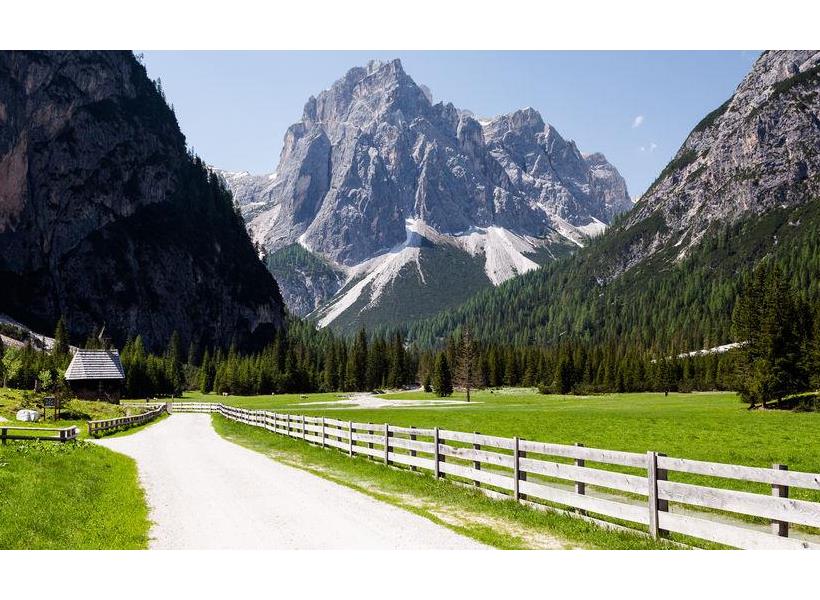 Ein breiter Wanderweg durch ein grünes Tal in den Dolomiten mit Wiesen, Wäldern und markanten Berggipfeln im Hintergrund.