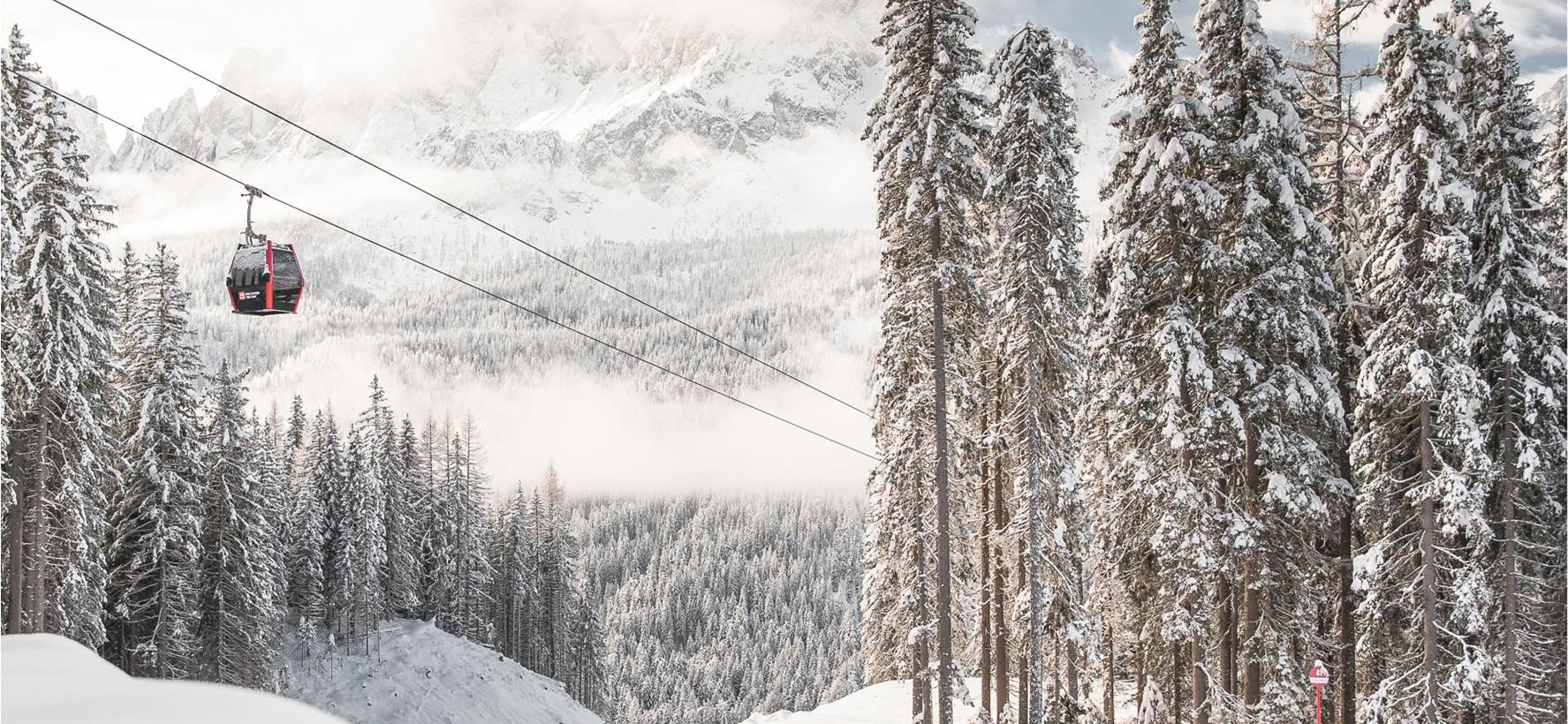 Rote Gondelbahn über verschneitem Winterwald in den Dolomiten mit schneebedeckten Berggipfeln im Hintergrund.