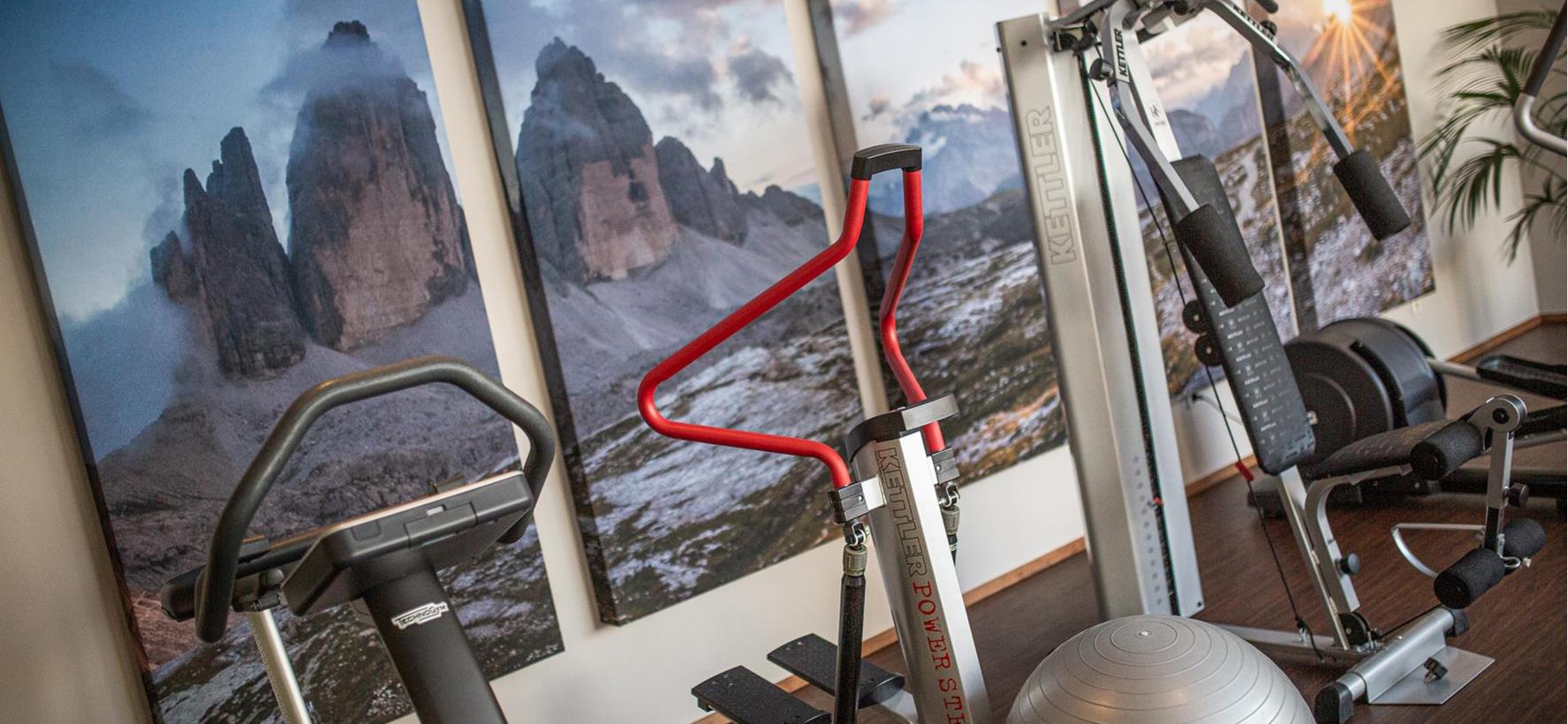 Fitnessraum mit verschiedenen Trainingsgeräten und großflächigem Drei-Zinnen-Motiv an der Wand.