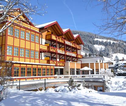 Hotel Villa Stefania im Winter mit schneebedecktem Garten davor und Wäldern im Hintergrund.