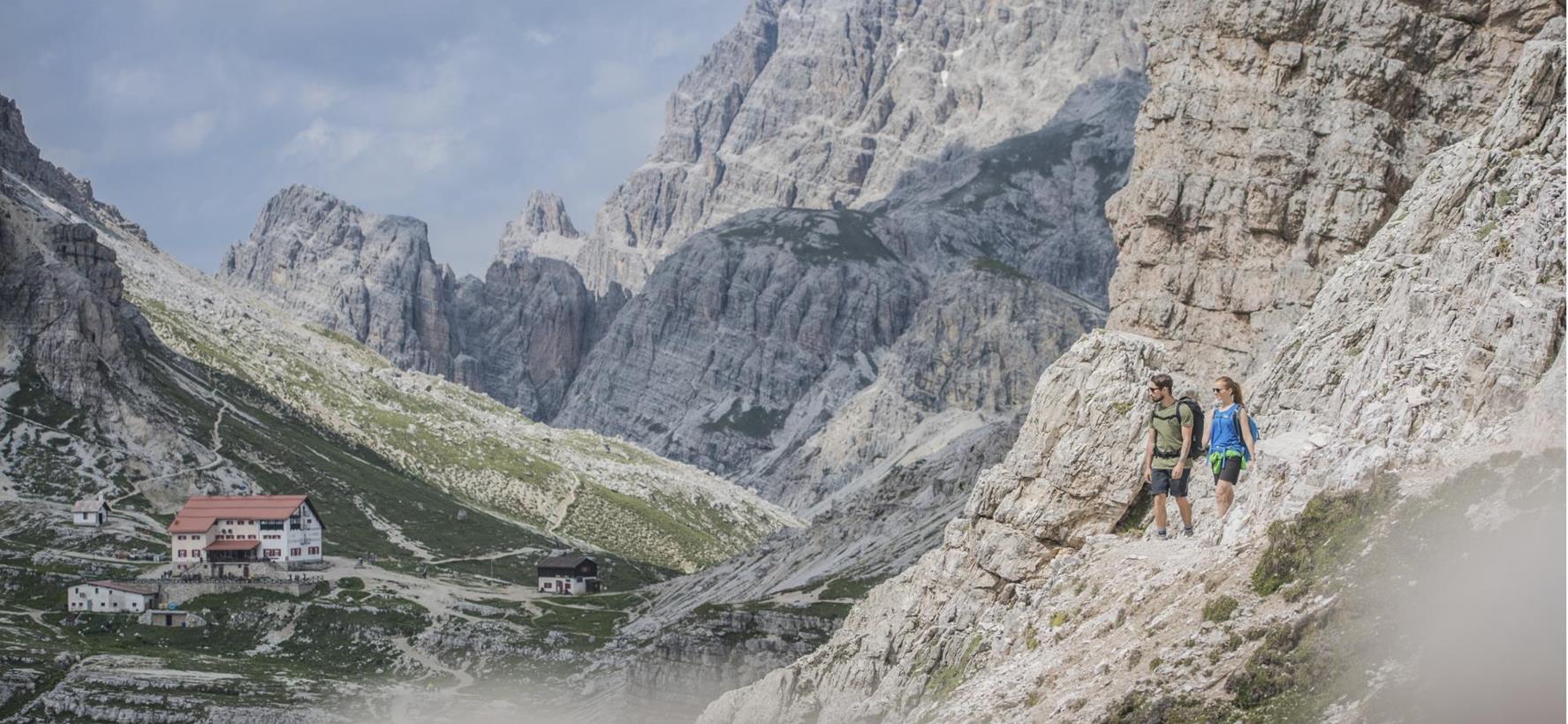 Two hikers on a rocky mountain trail amid the impressive Dolomite landscape around the Three Peaks of Lavaredo, with a mountain hut in the background.