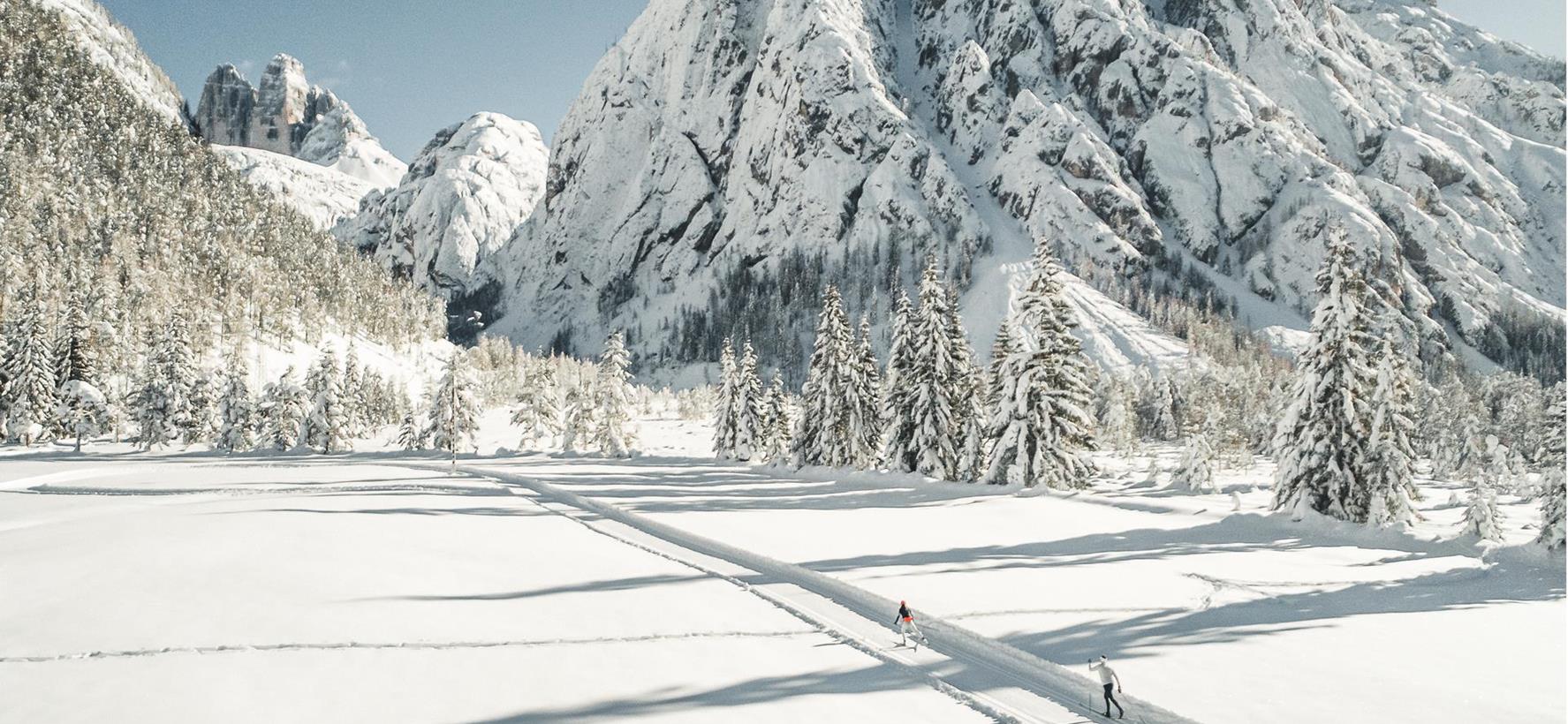 Langläufer auf einer Loipe in schneebedeckter Landschaft mit Bäumen und Gebirge im Hintergrund.