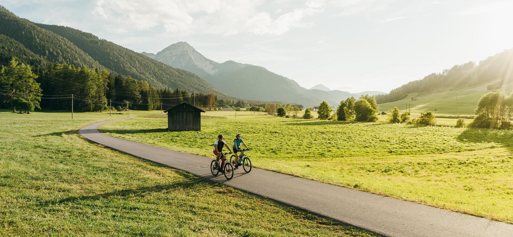 Two cyclists on a mountain road through the green South Tyrolean landscape, with forests and mountains in the background