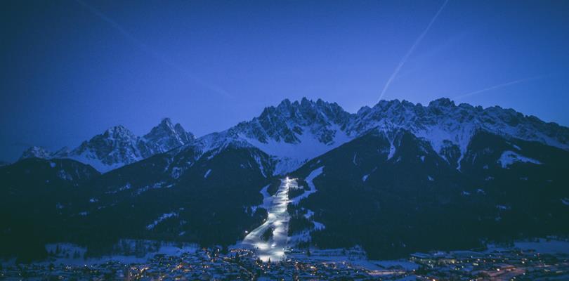 Night skiing in the Dolomites with an illuminated ski slope, skiers on the snow and a mountain landscape in the background.