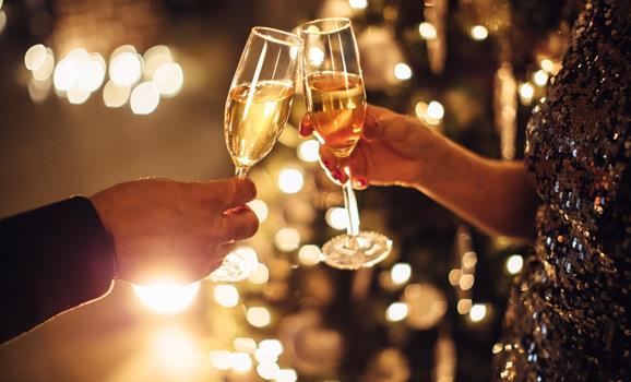 Two people toast with glasses of sparkling wine against a festively lit background.