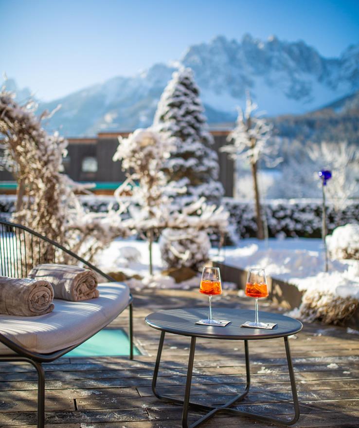 Orangefarbenes Getränk auf einem Tisch im verschneiten Garten der Villa Stefania mit Dolomitengipfeln im Hintergrund.