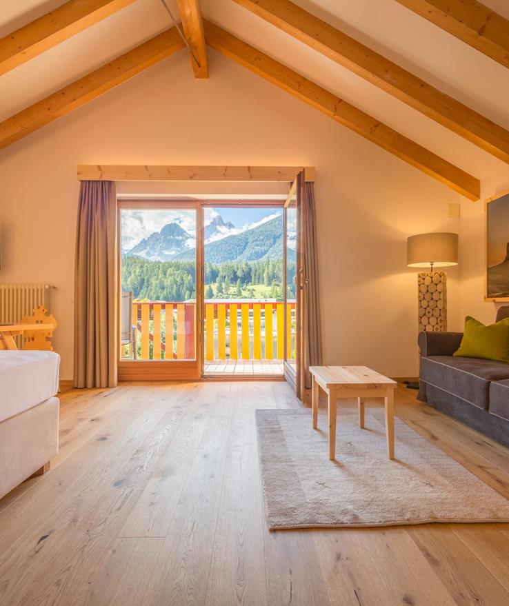 Living area of the Natura Suite with cosy sofa, bed, wooden beams on the ceiling and balcony with views of the Dolomites.