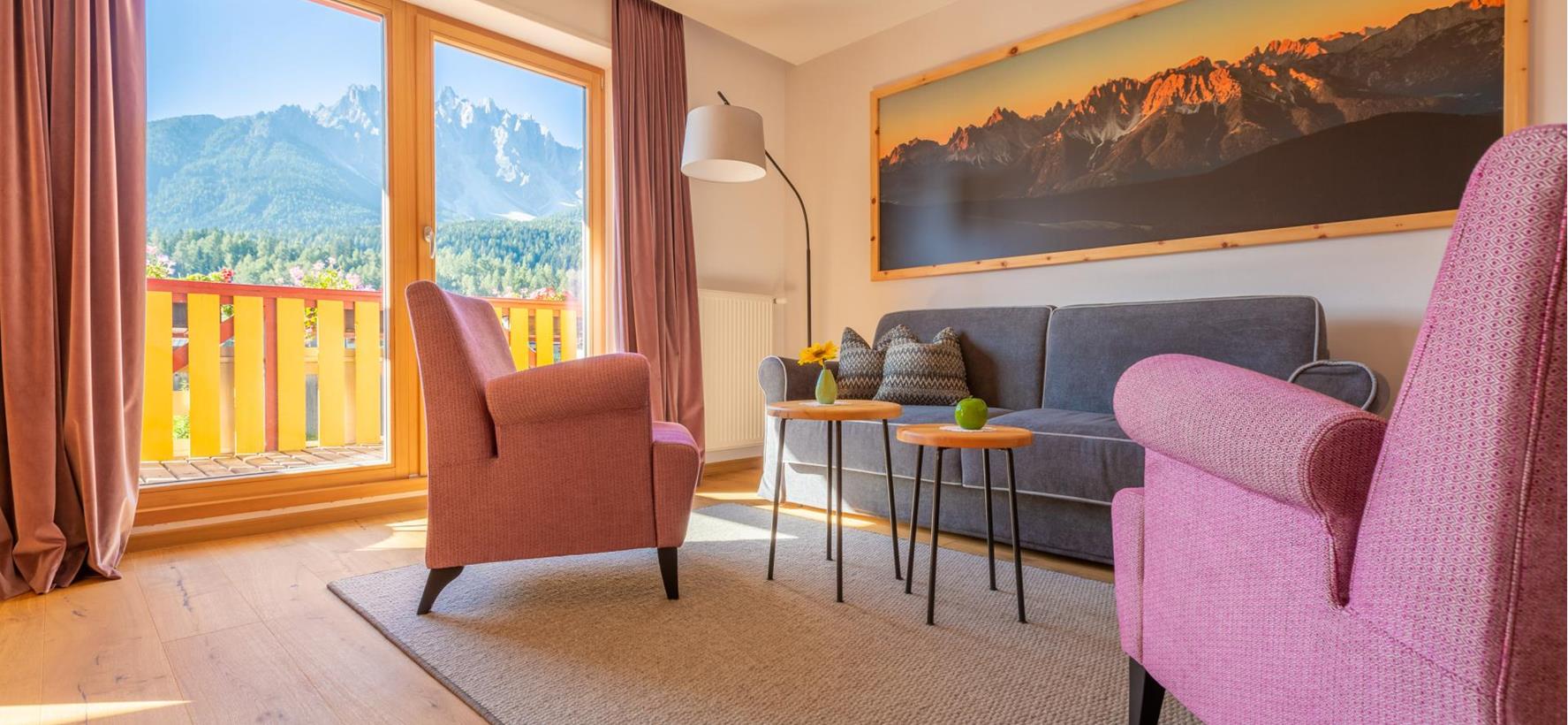 Sitzecke der Suite Dolomiti mit grauem Sofa, orangen Sesseln und Blick auf den Balkon und die Dolomiten.