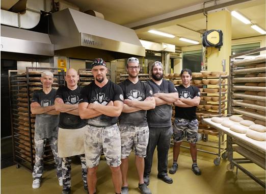 The Walder Bakery team, wearing work clothes, are standing together in the bakery in front of racks with baking trays.