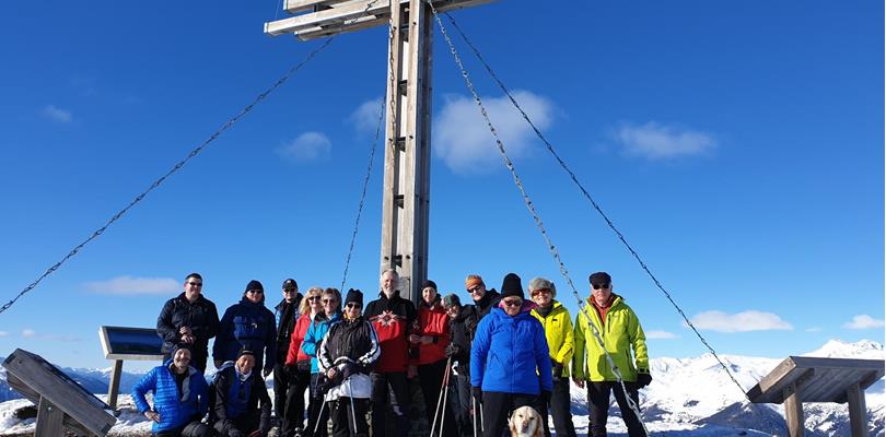 Hikers standing by a summit cross in a snow-covered landscape, with blue sky in the background.