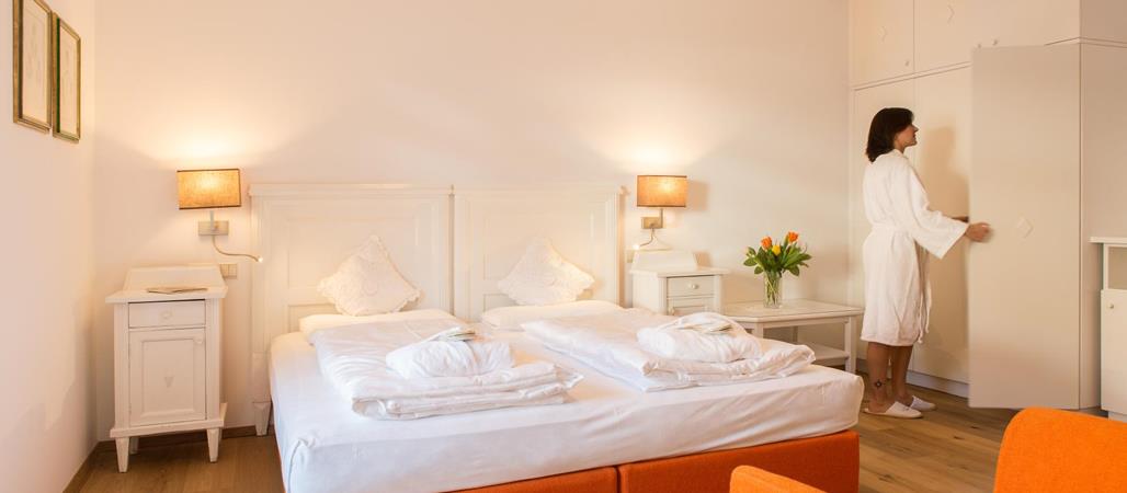 Classic double room with white linens, wall lamps, orange armchairs and a person in the background.