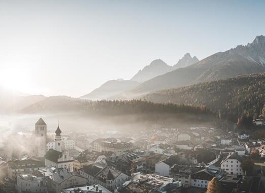 Luftaufnahme von Innichen im Herbst mit Wäldern und den massiven Bergen der Dolomiten im Hintergrund.