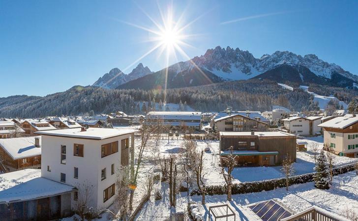 Vista panoramica sulle Dolomiti con paesaggio montano innevato, abitazioni e splendido sole.