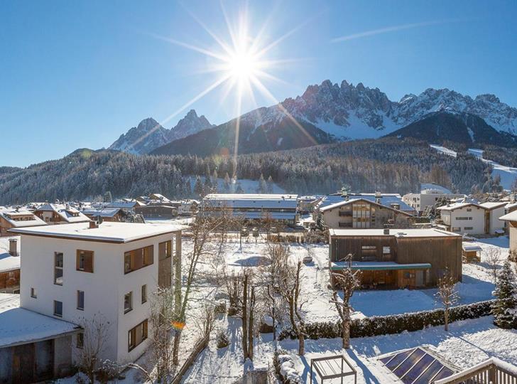 Vista panoramica sulle Dolomiti con paesaggio montano innevato, abitazioni e splendido sole.