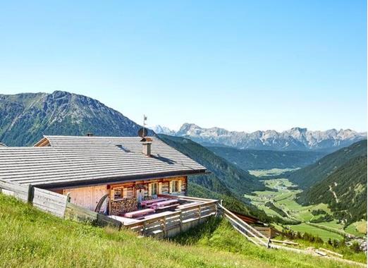 Traditional mountain hut on an alpine meadow overlooking a green valley and the surrounding mountain landscape.