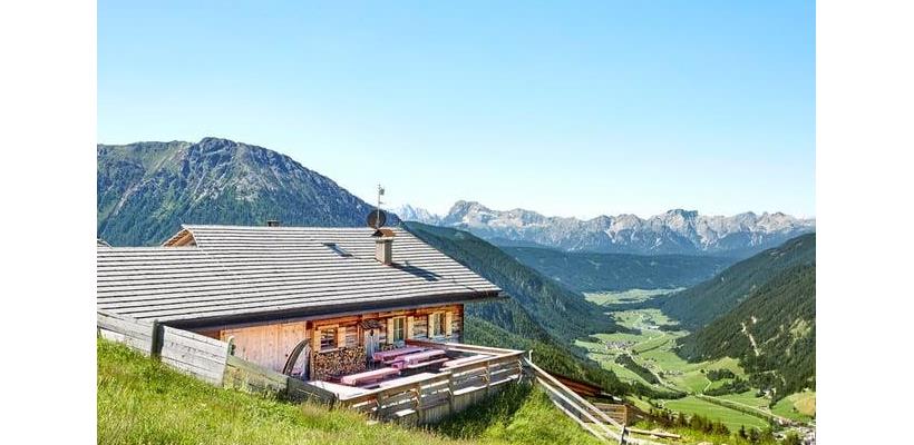 Traditional mountain hut on an alpine meadow overlooking a green valley and the surrounding mountain landscape.