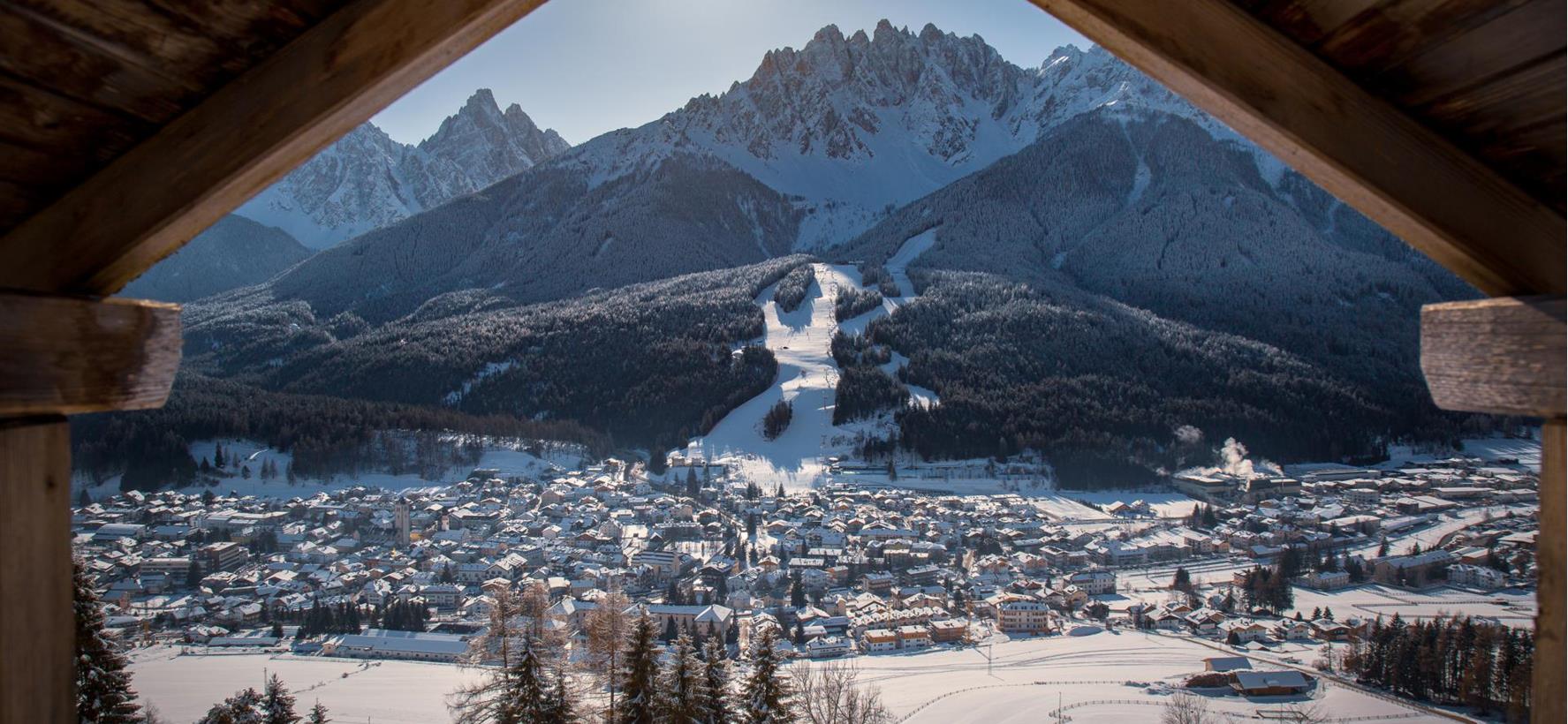 Vista invernale su San Candido con prati e boschi innevati e le suggestive cime delle Dolomiti sullo sfondo.