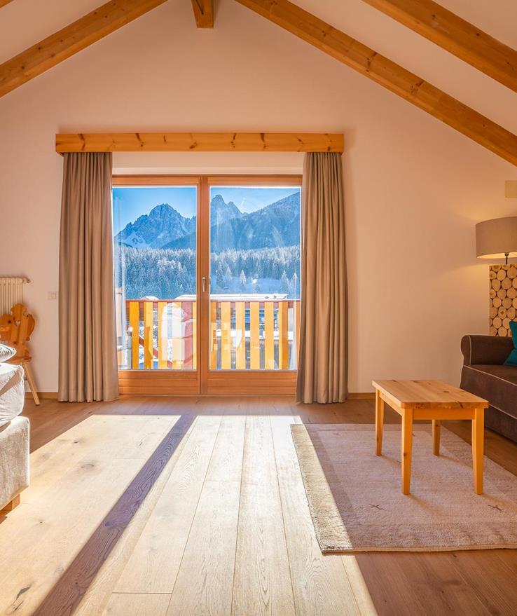 Living area of the Natura Suite with sofa, exposed wooden beams and balcony with panoramic views of the Dolomites.