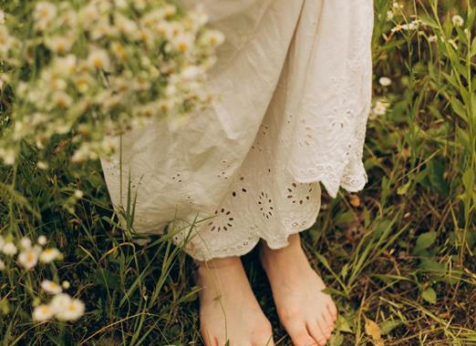 Persona a piedi nudi con un abito bianco in piedi su un prato fiorito con erba e piccoli fiori selvatici nella natura.