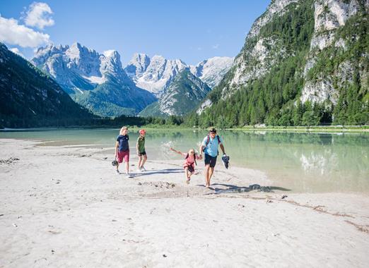 Familie am Ufer eines Bergsees in den Dolomiten mit klarem Wasser, grünen Wäldern und markanten Berggipfeln im Hintergrund.