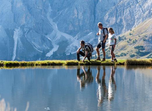 Gebirgssee im Vordergrund davor stehen Wanderer mit felsigem Gebirge im Hintergrund.