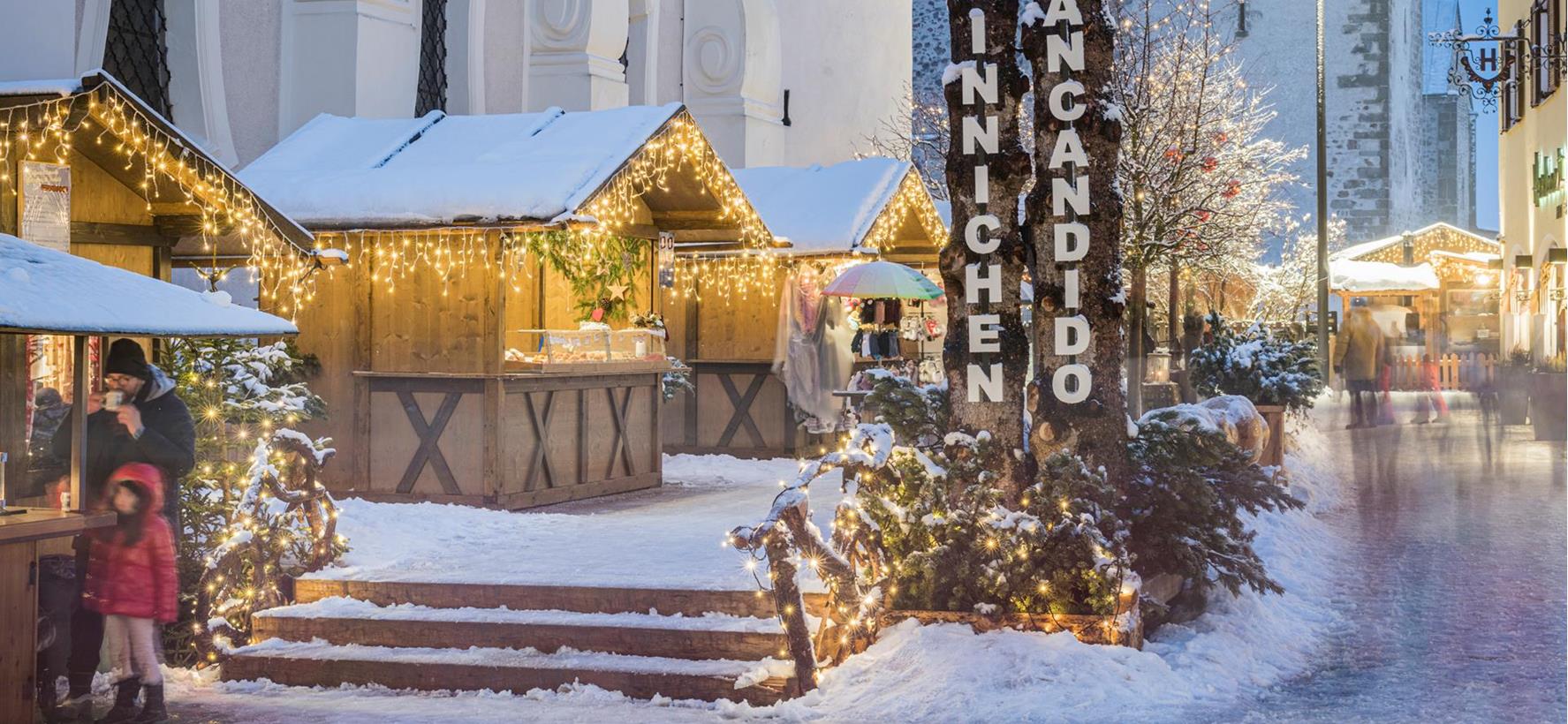 Verschneiter Platz in Innichen mit Weihnachtsmarkt, geschmücktem Weihnachtsbaum und festlich beleuchteter Kirche im Hintergrund.