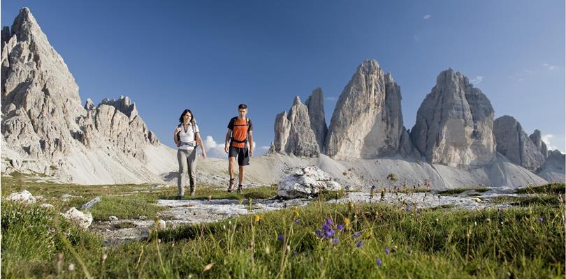 An alpine meadow in the foreground with two hikers and the Three Peaks of Lavaredo in the background.