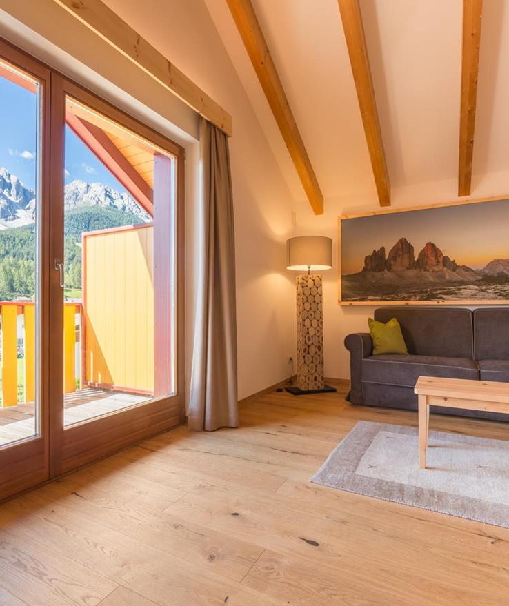 Living area of the Natura Suite at Hotel Villa Stefania with sofa, exposed wooden beams, light wooden floor and balcony with views of the Dolomites.