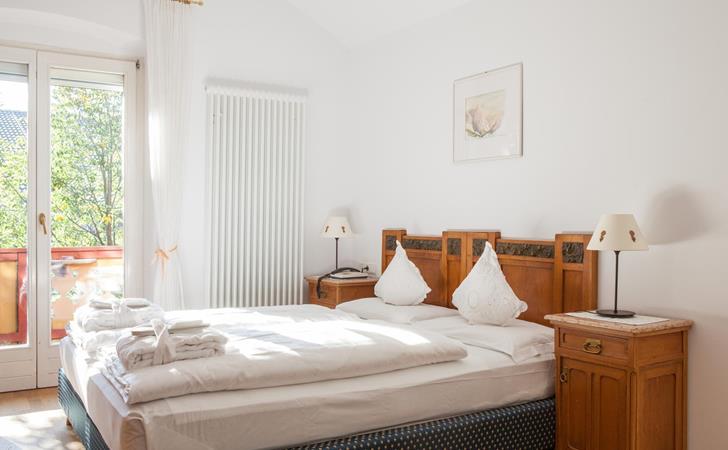 Classic double room with a wooden bed, white linens, bedside tables with reading lamps and a balcony door looking out onto greenery.