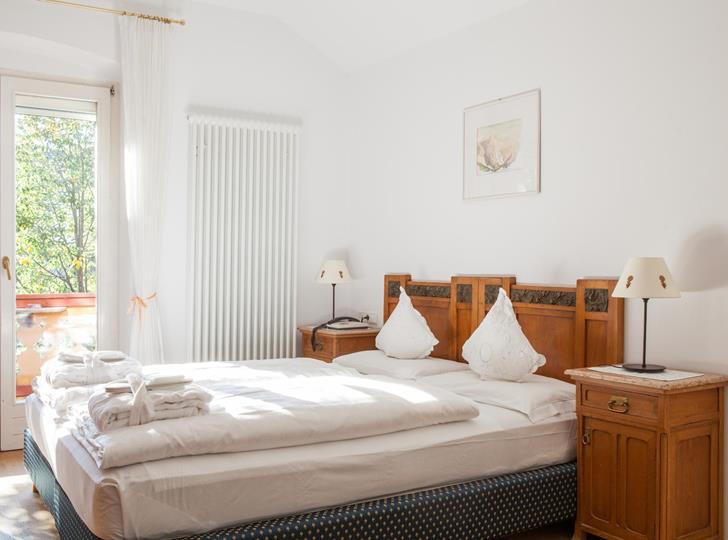 Doppelzimmer Classic mit Holzbett, weißer Bettwäsche, Nachtkästchen mit Leselampen und Balkontür mit Blick ins Grüne.