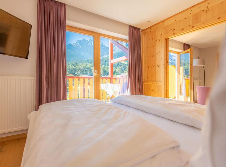 Suite Dolomiti mit Doppelbett, Flachbildfernseher, großer Fensterfront und Balkon mit Blick auf die Dolomiten.