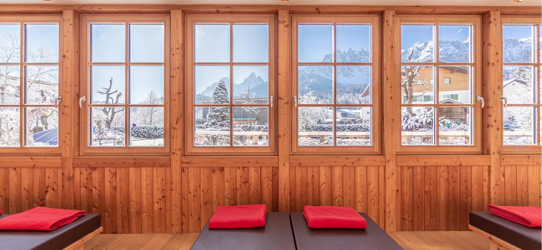 Sala relax con lettini, pareti in legno e ampie finestre panoramiche con vista sul paesaggio montano.