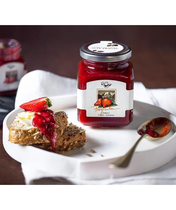 Jar of Alpe Pragas strawberry jam on a plate with bread, butter, strawberry jam and a fresh strawberry.