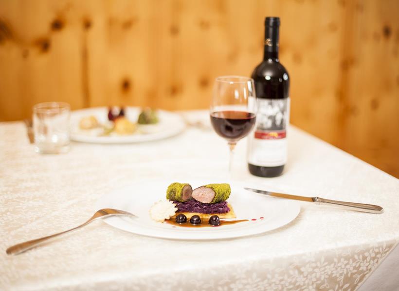 Tavolo apparecchiato con piatto principale, bicchiere di vino rosso e bottiglia di vino sullo sfondo.