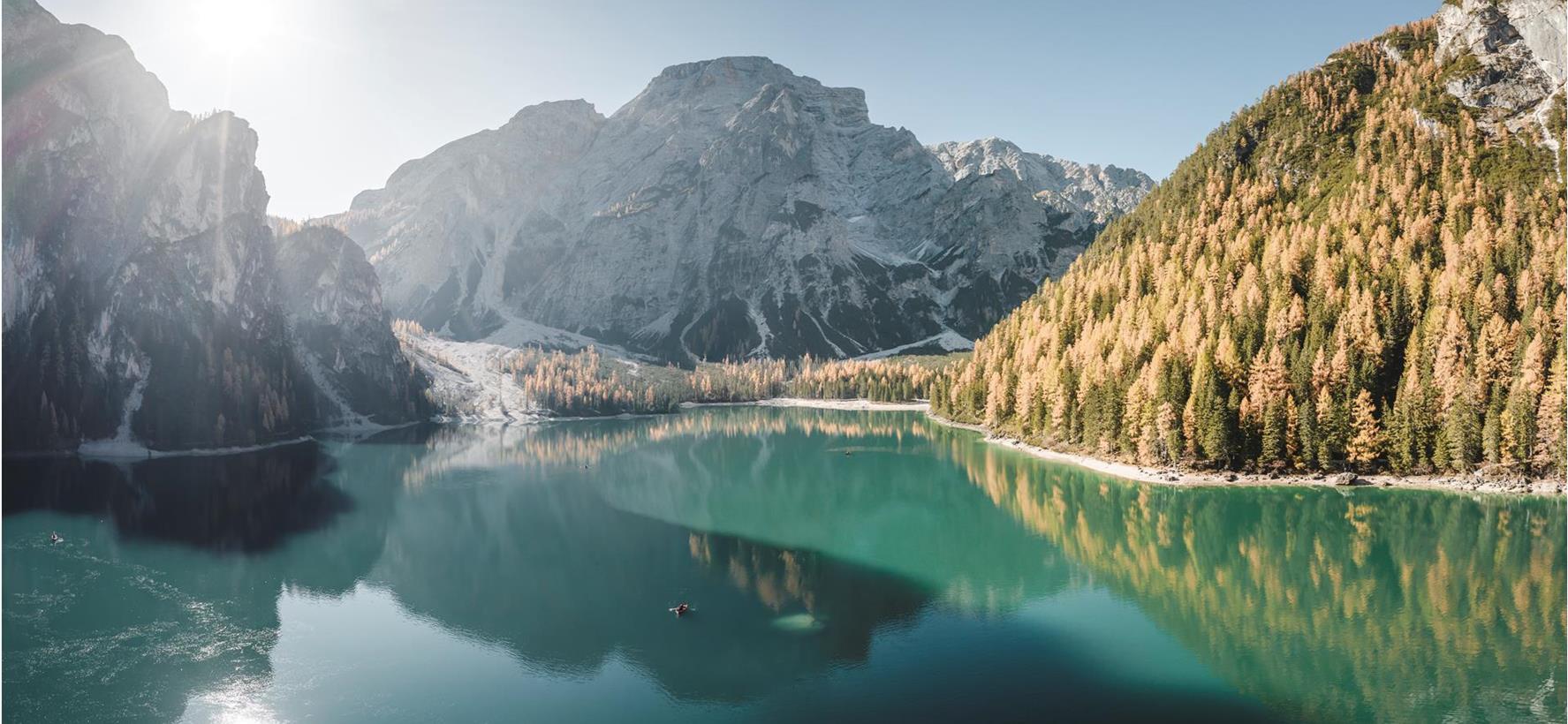 Der Pragser Wildsee im Sonnenschein mit herbstlich gefärbten Bäumen und markanten Bergen im Hintergrund.