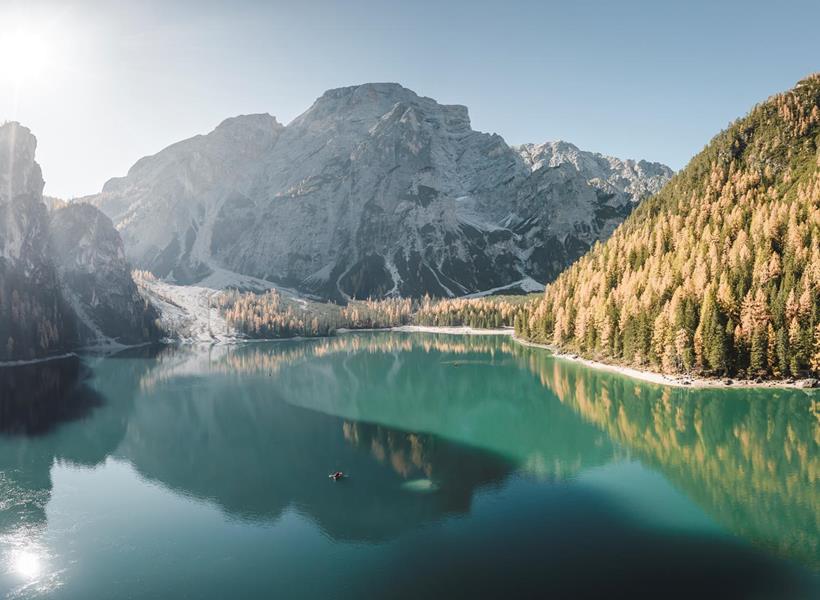 Der Pragser Wildsee im Sonnenschein mit herbstlich gefärbten Bäumen und markanten Bergen im Hintergrund.