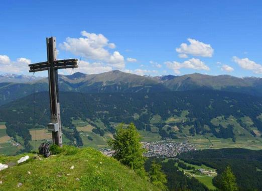 Wooden cross on the Gantraste with a view of a vast alpine landscape, valleys and surrounding mountains under a blue sky.