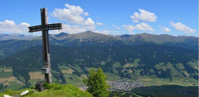 Croce in legno sulla Gantraste con vista su un ampio paesaggio alpino, vallate e montagne circostanti sotto il cielo azzurro.