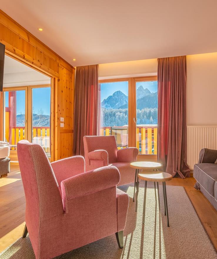 Sitzecke der Suite Dolomiti mit grauem Sofa, orangen Sesseln und Blick auf den Balkon und die Dolomiten.