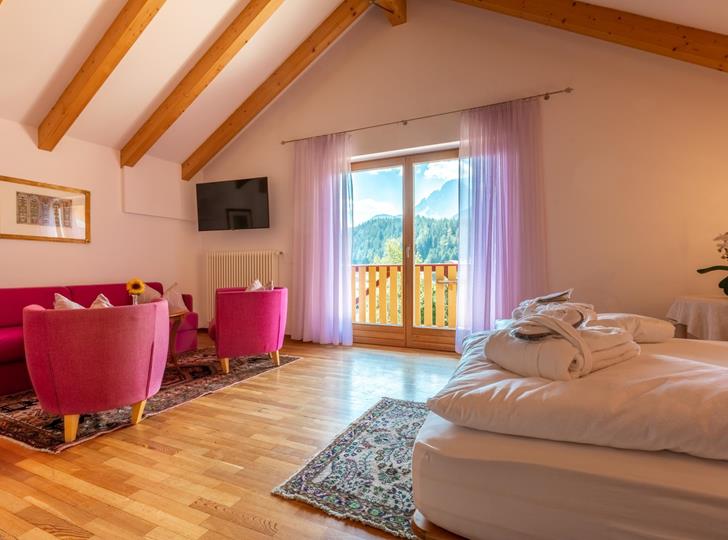 Junior Suite con letto matrimoniale, angolo soggiorno, pavimento in legno chiaro e balcone con vista sulle Dolomiti.
