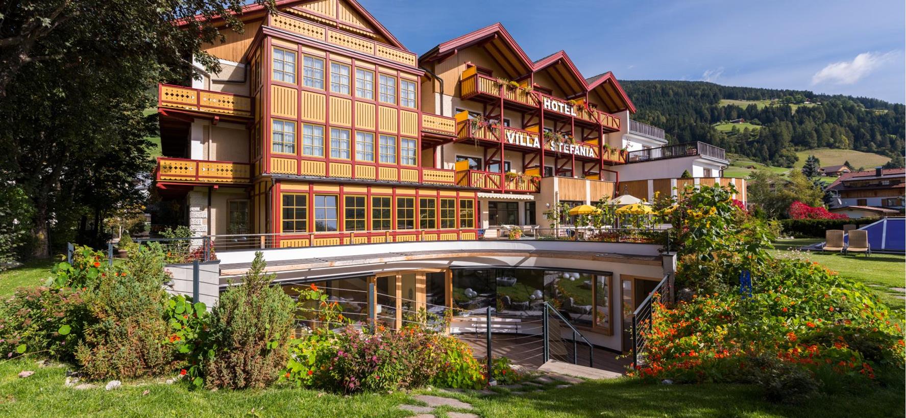 Hotel Villa Stefania in Innichen mit Garten, Bäumen und Wäldern im Hintergrund.