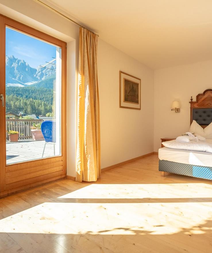 Junior Suite bedroom with a double bed and a panoramic window overlooking the terrace and the Dolomites.